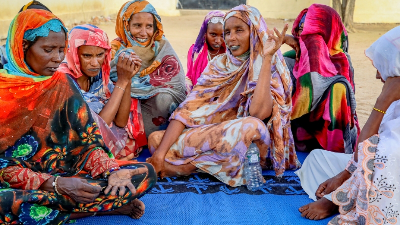 Ehemalige Fistelbetroffene finden zu neuer Stärke Frauen in bunten Kleidern sitzen nebeneinander. Eine hebt die Hand, um etwas zu sagen.