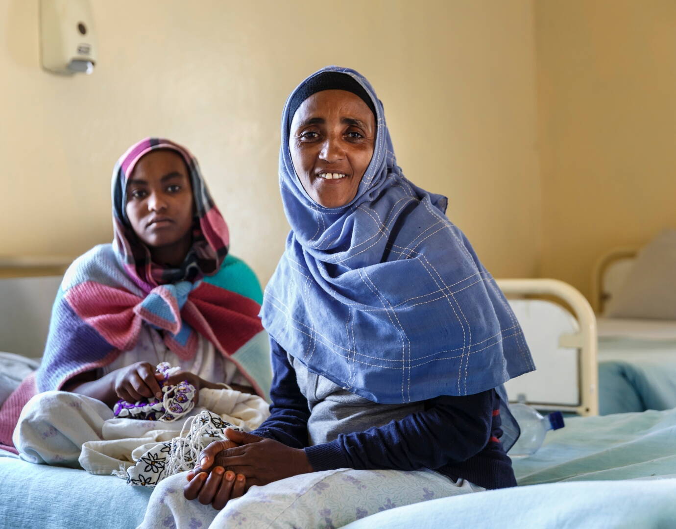 Jetzt spenden! Zwei Frauen sitzen auf einem Spitalbett und schauen in die Kamera.